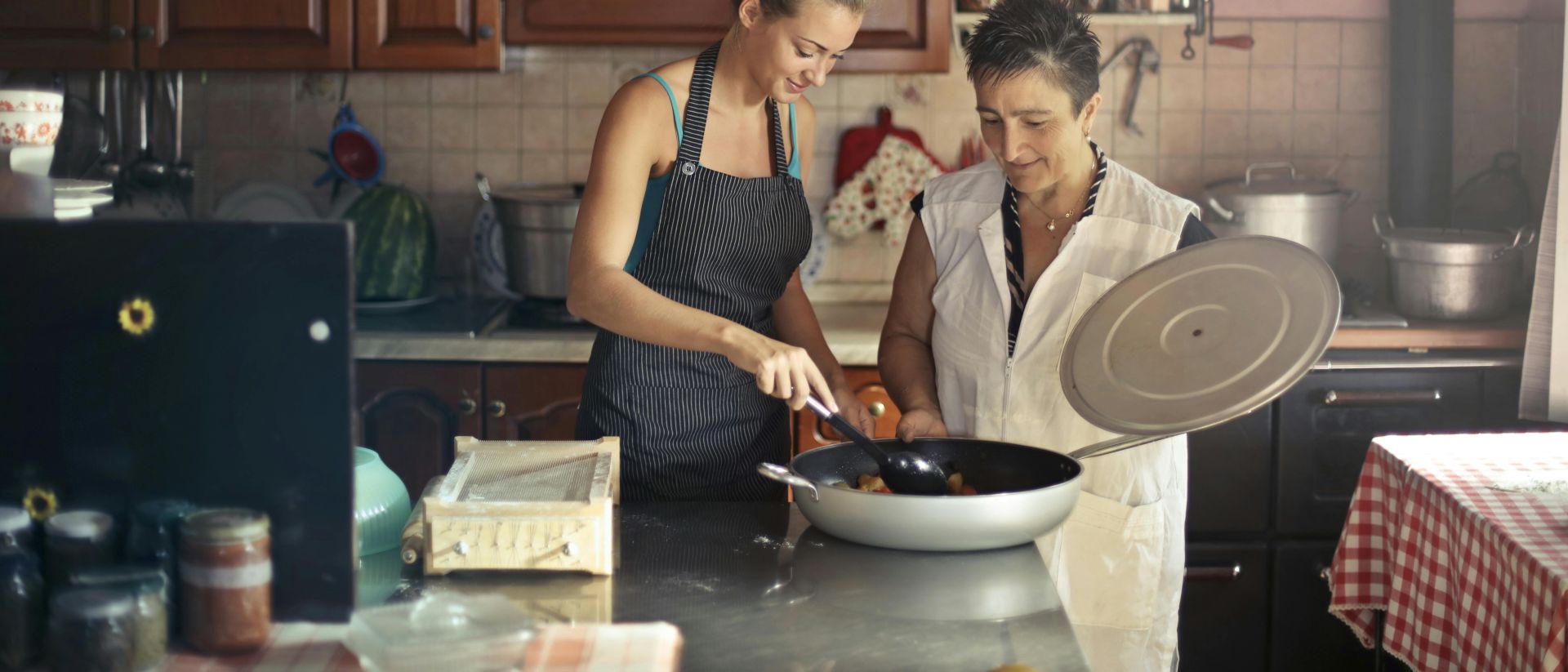 Daughter and senior mother standing at table in kitchen and stirring dish in frying pan while preparing food for dinner